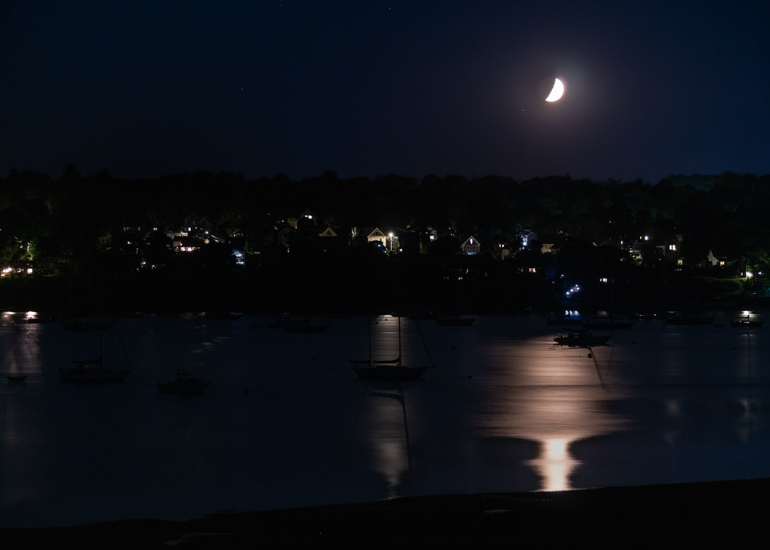 The moon hovers above a hill over a town, it's light and the towns light reflected in the bay